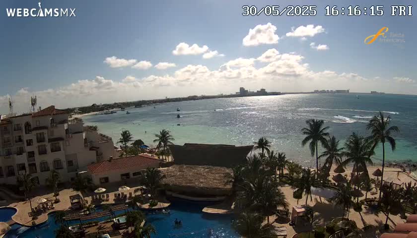 A partly sunny day shows a resort with multiple pools overlooking a calm ocean with a beach and palm trees.