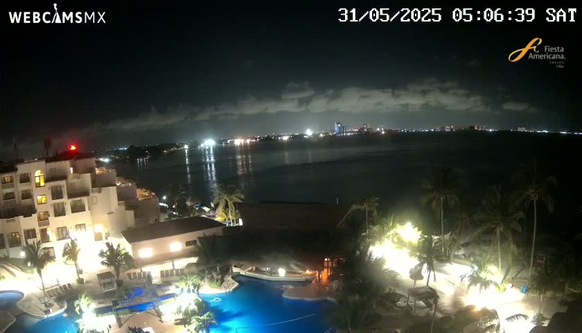 A nighttime view of a resort with swimming pools, palm trees, and a city skyline across the water under a partly cloudy sky.
