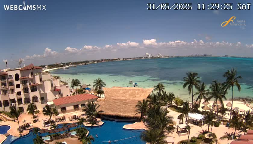 An aerial view shows a resort with swimming pools and palm trees on a sunny day next to a calm, turquoise ocean with a beach and distant coastline.