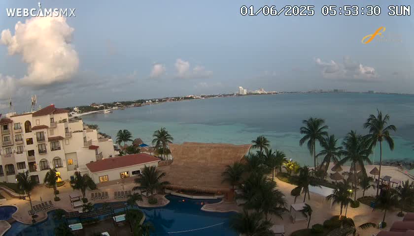 An aerial view of a resort on a tropical beach at dawn, with partly cloudy skies and calm ocean water.