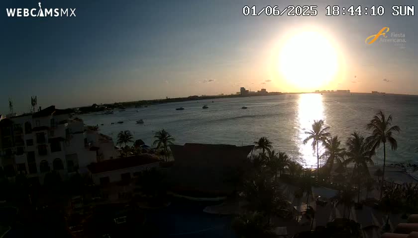 A coastal resort town at sunset, with a bright sun reflecting on calm ocean water, palm trees silhouetted against the light, and a multi-story building in the foreground.