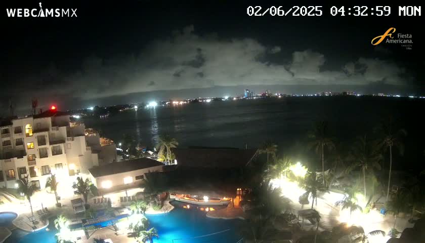 A nighttime view of a resort complex with swimming pools, illuminated buildings, and palm trees overlooking a calm body of water with a city skyline in the distance under a partly cloudy sky.
