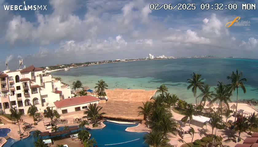 A partly cloudy day reveals a beachfront resort with palm trees, swimming pools, and a thatch-roofed structure, overlooking a calm turquoise ocean and a distant cityscape.