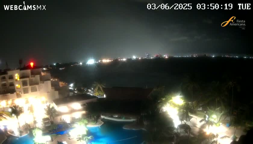 A nighttime view of a resort complex with illuminated buildings and pools overlooking a dark body of water and a distant city skyline under a cloudy sky.