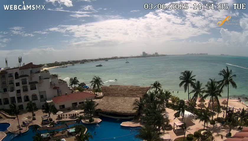 An aerial view of a resort overlooking a calm, partly cloudy ocean with palm trees lining the beach.