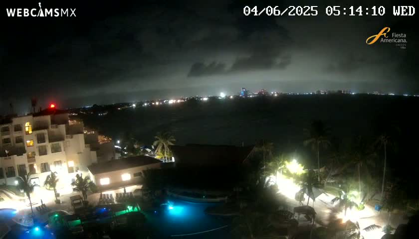 A nighttime view shows a resort with illuminated pools and buildings overlooking a dark body of water with a city skyline faintly visible on the horizon under an overcast sky.