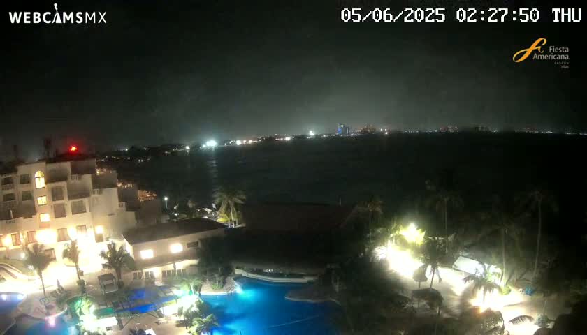 A nighttime view of a resort complex with illuminated buildings, pools, and palm trees overlooking a dark body of water under an overcast sky.
