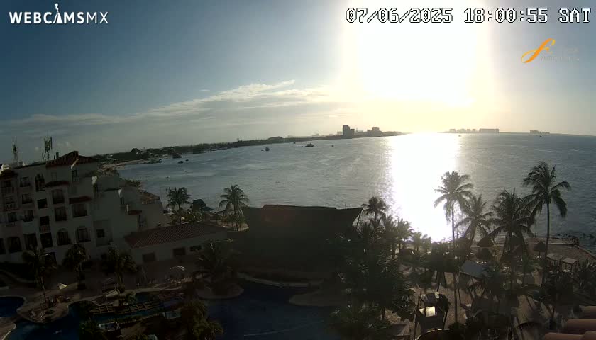 A partly sunny sunset over a calm ocean, viewed from a high vantage point overlooking a resort with pools and palm trees.