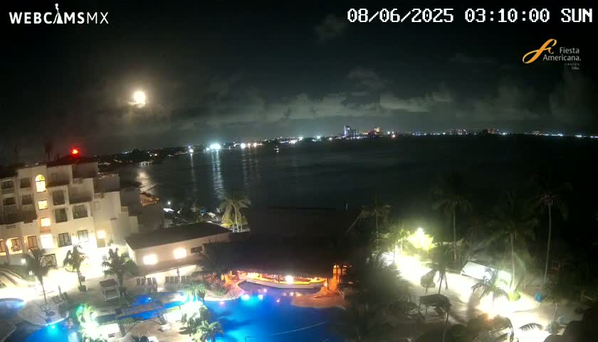 A brightly lit resort complex with swimming pools is shown at night under a partly cloudy sky and a full moon, overlooking a calm body of water with a city skyline in the distance.