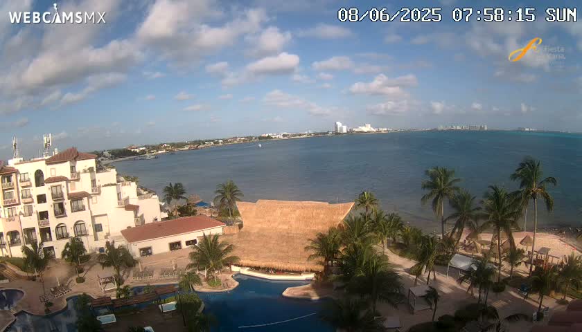 A partly cloudy sunny day overlooks a resort with a pool and thatch-roofed structure, situated near a calm ocean with a distant cityscape visible on the horizon.