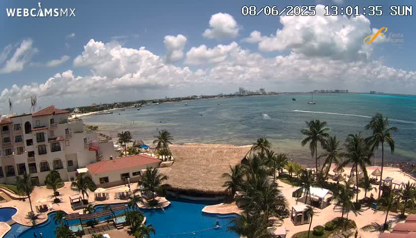 An aerial view of a resort on a sunny day with partly cloudy skies, showing a beachfront property with multiple pools and palm trees, overlooking a calm ocean with a distant shoreline.