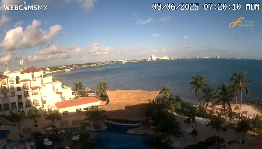A partly cloudy morning scene overlooks a resort with pools and palm trees bordering a calm ocean with a distant city skyline visible.