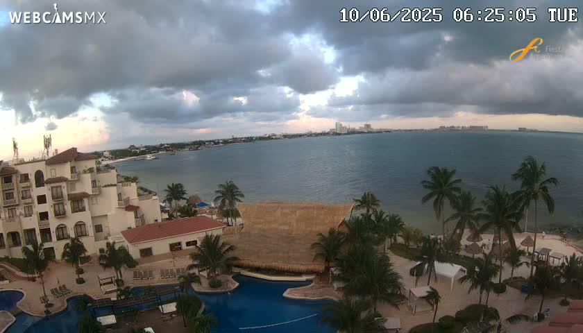 A resort with a large pool overlooks a calm ocean under a partly cloudy sky.
