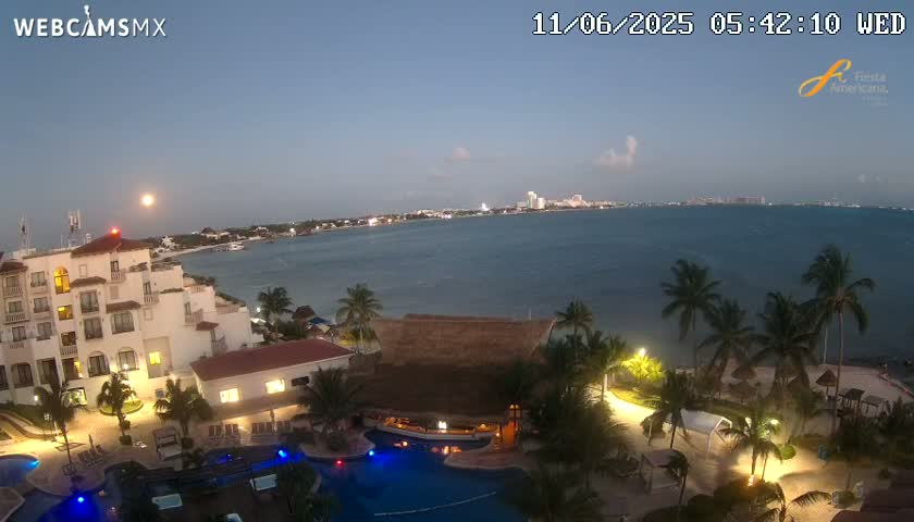 A brightly lit resort with a swimming pool overlooks a calm, dark-blue ocean at dusk, under a mostly clear sky.