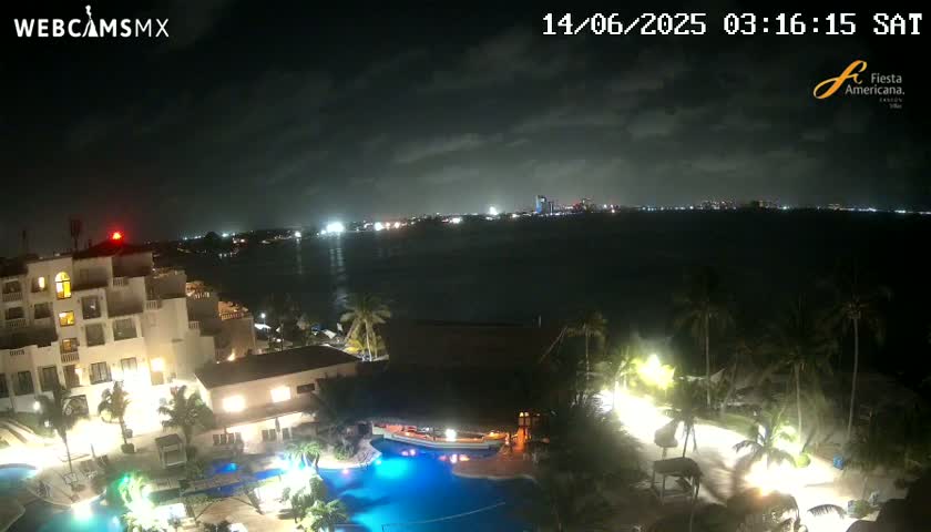 A nighttime view of a resort with pools, palm trees, and buildings overlooking a dark body of water with city lights in the distance under a partly cloudy sky.
