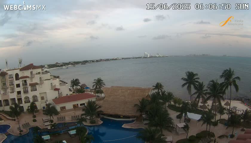 An aerial view shows a resort with palm trees, pools, and buildings overlooking a calm, overcast body of water with a city visible in the distance.