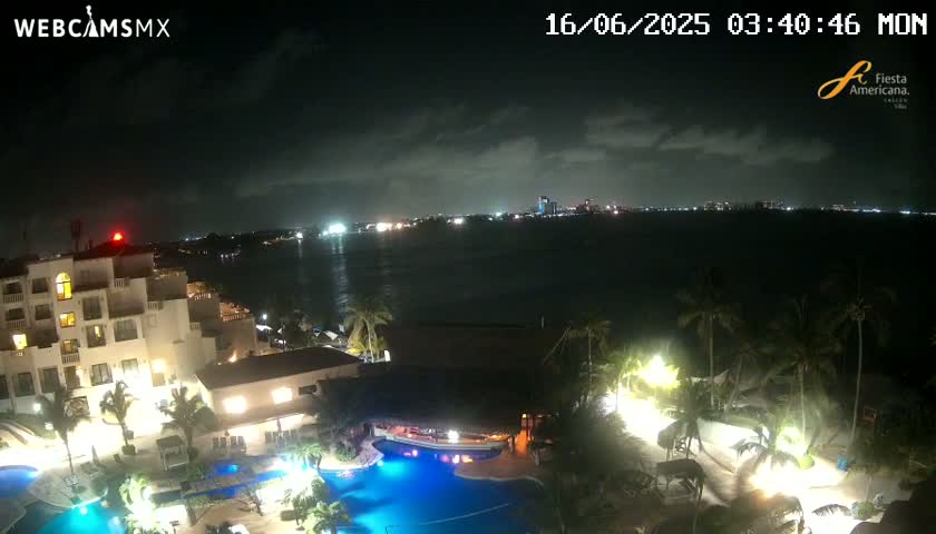 A nighttime view of a resort complex with swimming pools, illuminated buildings, and palm trees overlooking a calm, dark ocean under a partly cloudy sky.