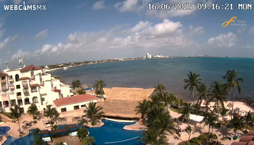 A resort with multiple pools and palm trees sits on the coast, overlooking a calm ocean under a partly cloudy sky.