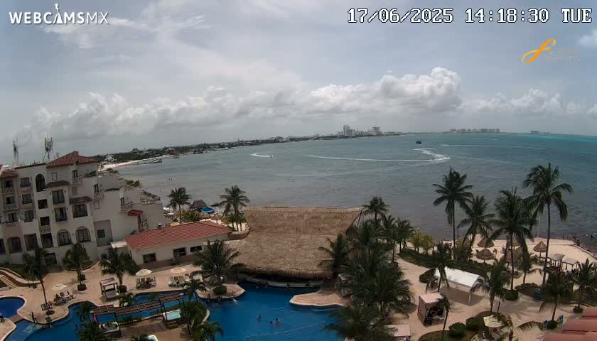 An aerial view of a resort on a coast with a partly cloudy sky shows a large swimming pool and a beach with palm trees, all overlooking a calm ocean.