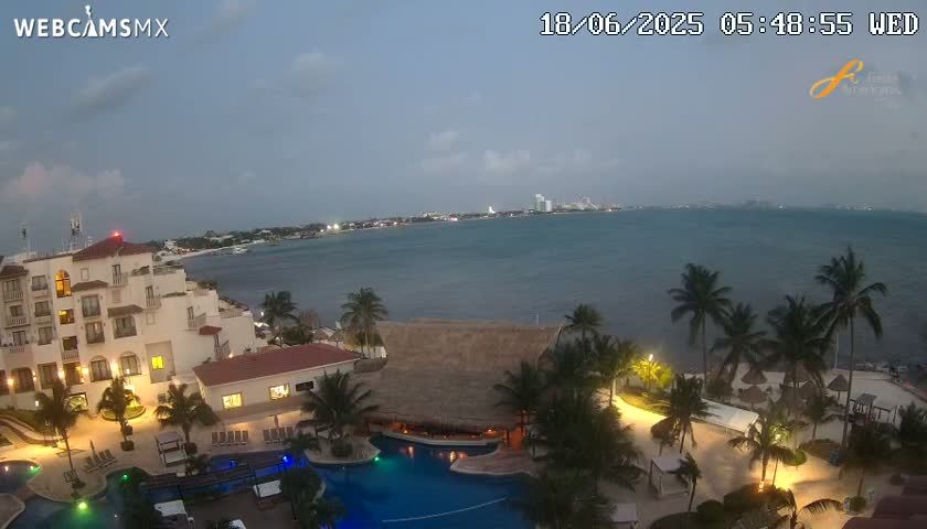An overcast evening scene shows a resort with pools and palm trees overlooking a calm body of water and a distant city skyline.