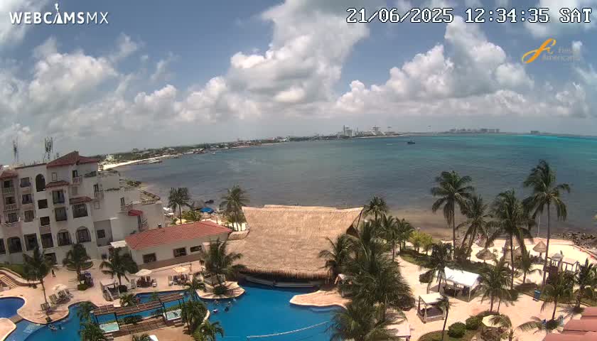 A partly cloudy day overlooks a resort with pools and palm trees beside a calm, turquoise ocean and a distant shoreline.