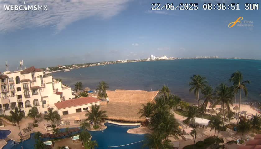 A resort with swimming pools and palm trees overlooks a calm ocean on a partly sunny day.