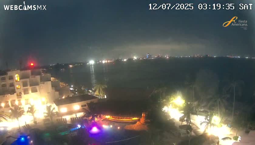 A nighttime view of a resort by the water, showing the resort's illuminated buildings and pools, with a city skyline visible across the dark water in the distance.