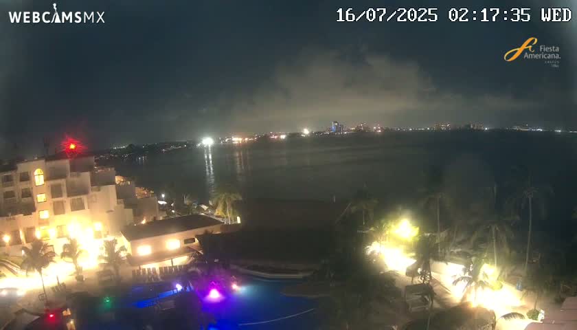 A nighttime view of a resort with a pool, illuminated by artificial lights, overlooking a calm body of water with a city skyline faintly visible in the distance under a partly cloudy sky.