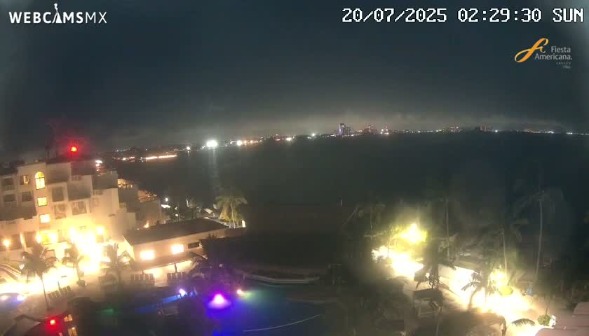 A nighttime view of a resort complex with a swimming pool, illuminated buildings, and palm trees, overlooking a calm body of water with distant city lights.
