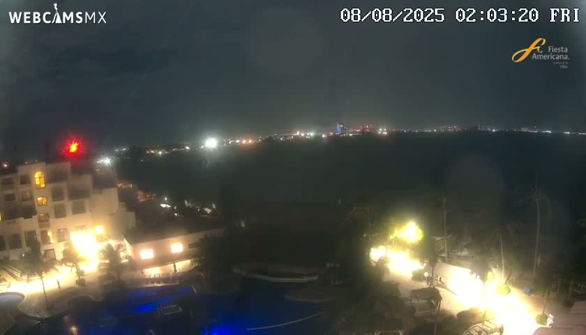 A nighttime view shows a resort complex with pools and palm trees, overlooking a dark body of water with a distant city skyline illuminated under a mostly clear sky.