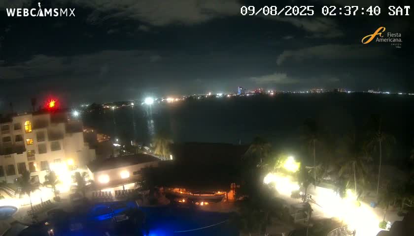 A nighttime view shows a resort with pools and palm trees, overlooking a calm body of water with a city skyline illuminated in the distance under a partly cloudy sky.