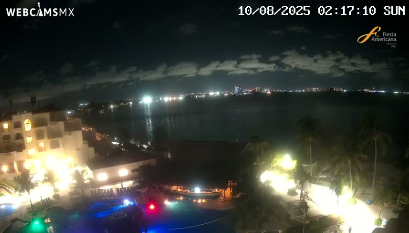 A nighttime view shows a resort complex with illuminated pools and palm trees, overlooking a calm body of water with a city skyline visible in the distance under a partly cloudy sky.