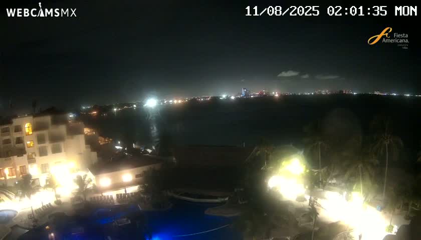 A nighttime view shows a resort with illuminated pools and buildings overlooking a dark body of water with a city skyline faintly visible in the distance under a mostly clear sky.