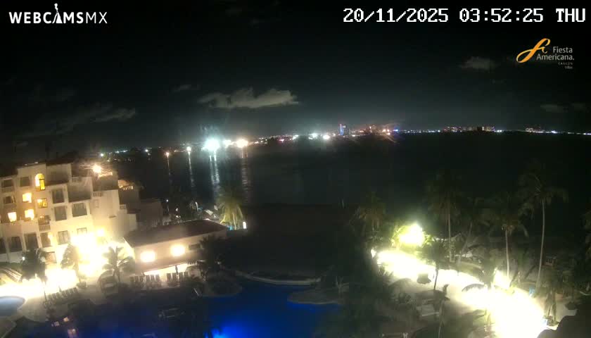 A nighttime view captures an illuminated resort with glowing pools and palm trees beside a calm body of water reflecting distant city lights under a dark, partly cloudy sky.