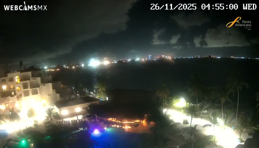 The image shows a cloudy night over a brightly lit hotel and pool area with palm trees, overlooking a dark body of water and a distant city skyline.