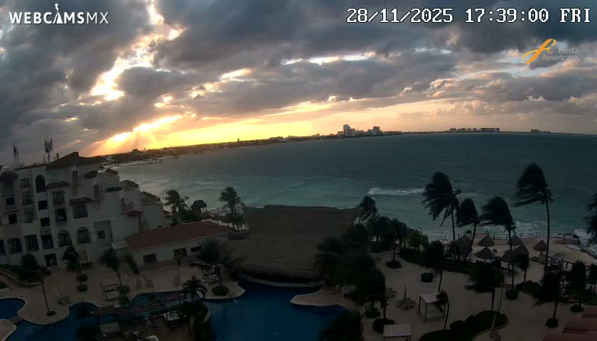 A coastal resort scene is captured at sunset under a heavily clouded sky with sunbeams, revealing choppy turquoise ocean waves and palm trees swaying in windy conditions, with a distant city along the horizon.