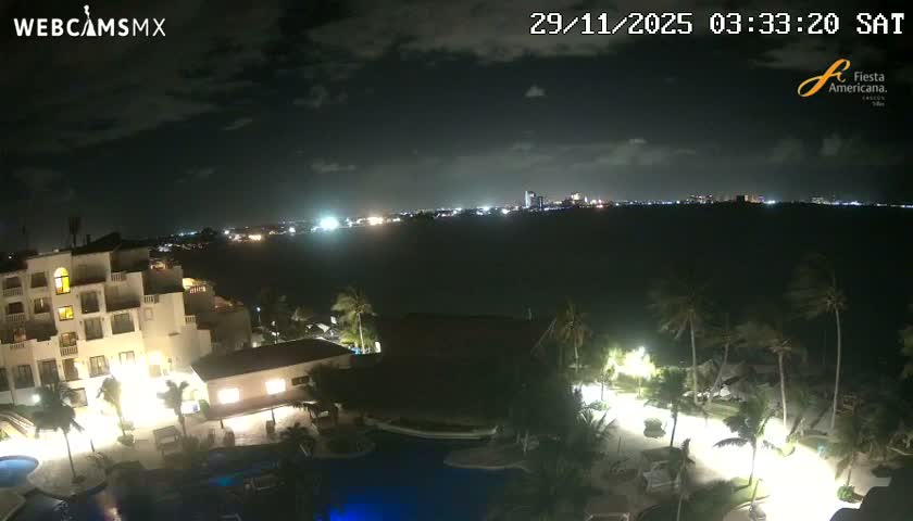 An elevated nighttime view reveals a brightly lit resort complex with pools and palm trees in the foreground, overlooking a dark body of water and an illuminated distant city skyline under a partly cloudy sky.