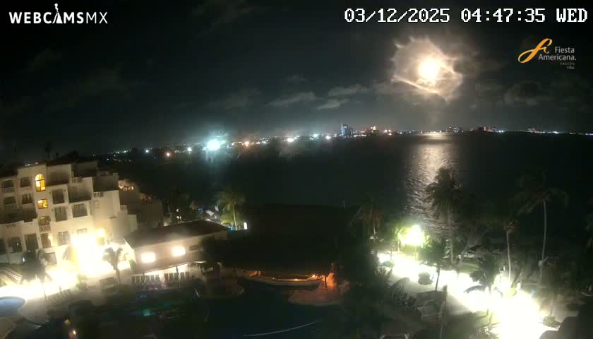 At night, a coastal scene reveals a brightly moonlit sky with scattered clouds above a calm body of water reflecting the moon and distant city lights, alongside an illuminated resort complex featuring buildings, pools, and palm trees.