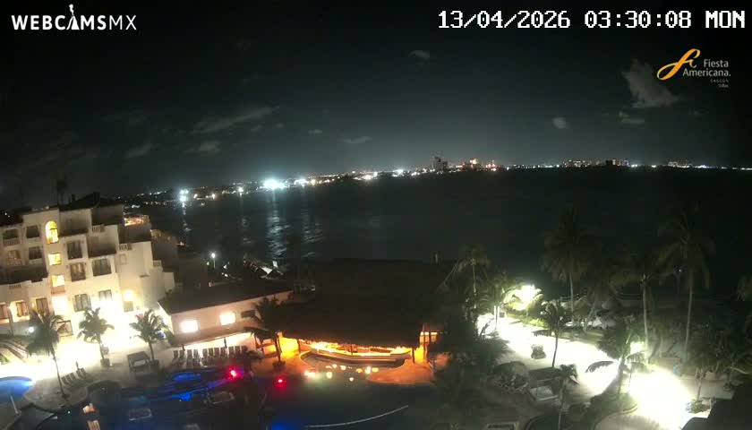 At night, a coastal scene reveals a brightly moonlit sky with scattered clouds above a calm body of water reflecting the moon and distant city lights, alongside an illuminated resort complex featuring buildings, pools, and palm trees.