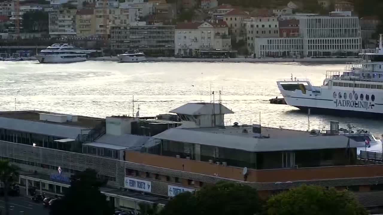 A ferry and other boats are docked in a harbor next to a building, with calm, reflective water under a mostly sunny sky.