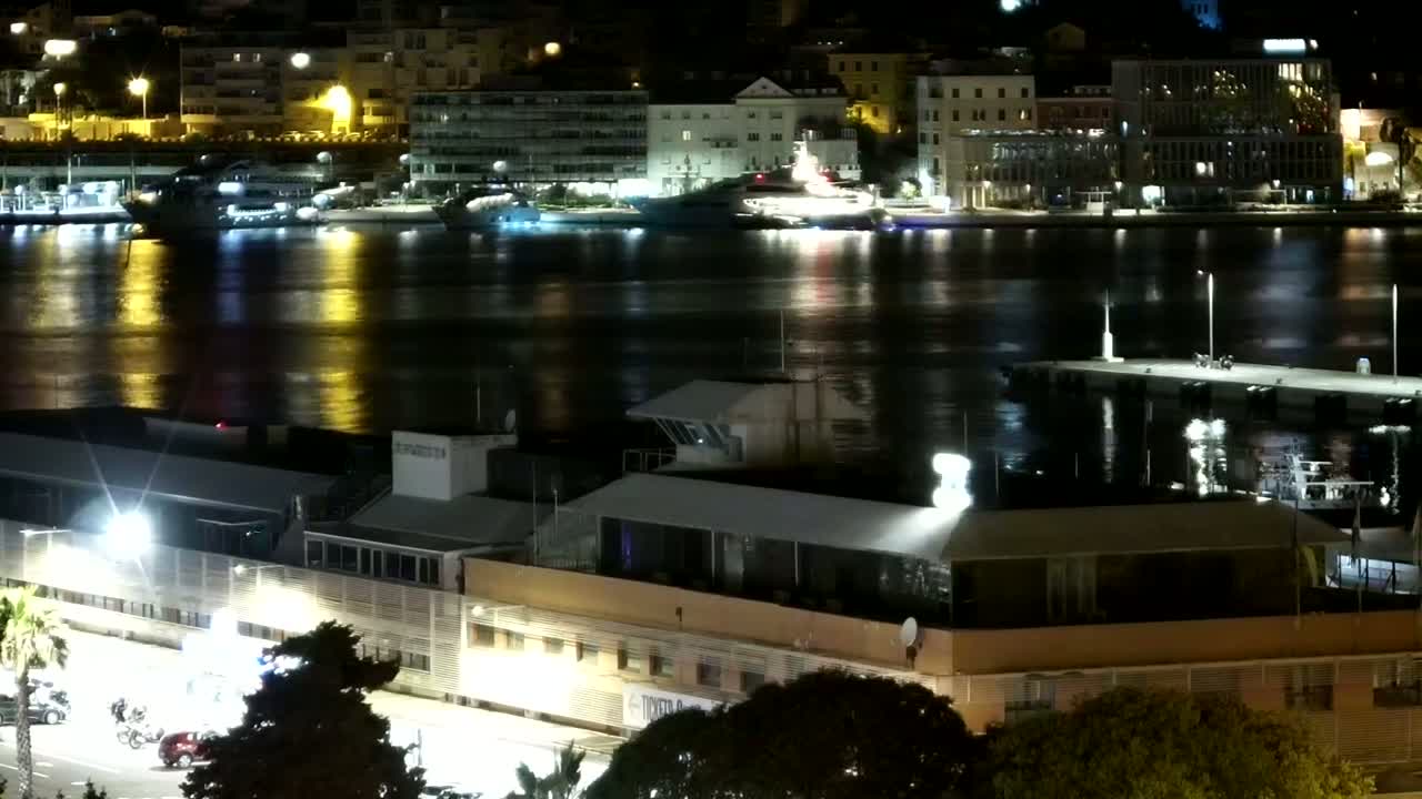 A nighttime view of a calm harbor reflecting city lights, with boats moored near a waterfront building.