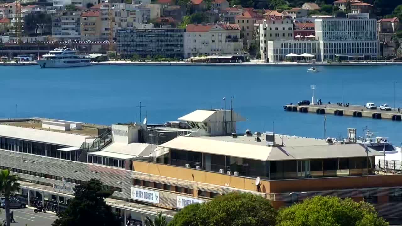 A ferry terminal building overlooks a calm blue harbor with a docked cruise ship and a city in the background under sunny skies.