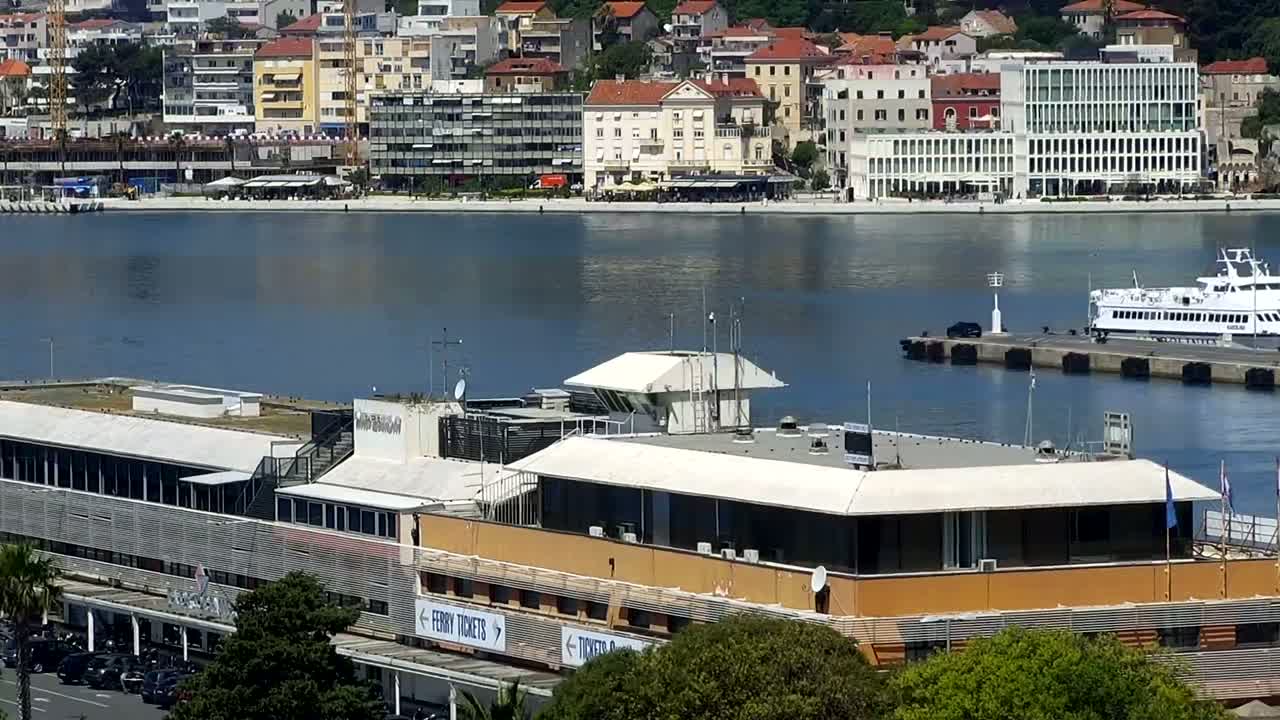 A ferry is docked at a port next to a large building with a flat roof, across a calm body of water from a town with many buildings.