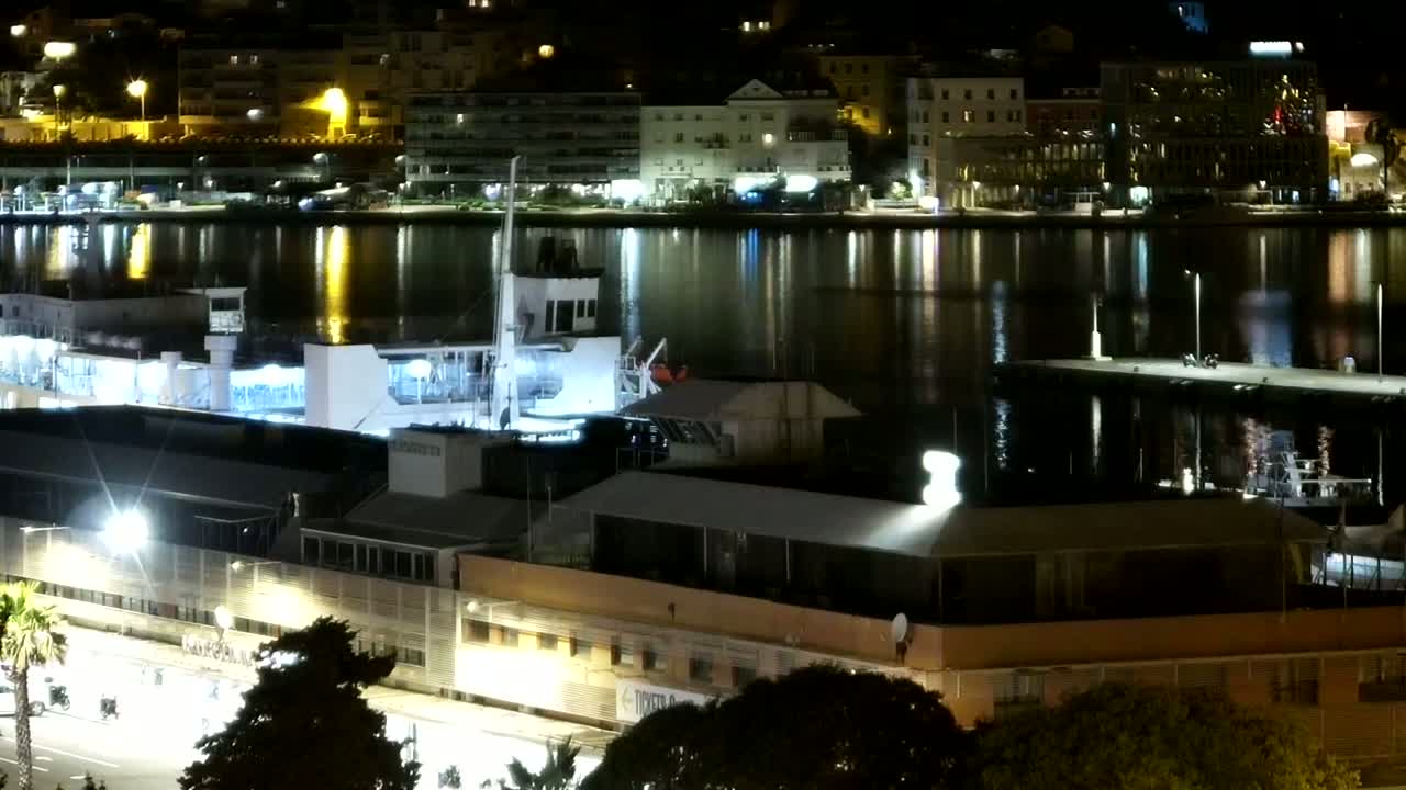 A nighttime view of a harbor with a large docked ship, illuminated buildings along the shore, and calm, reflective water.