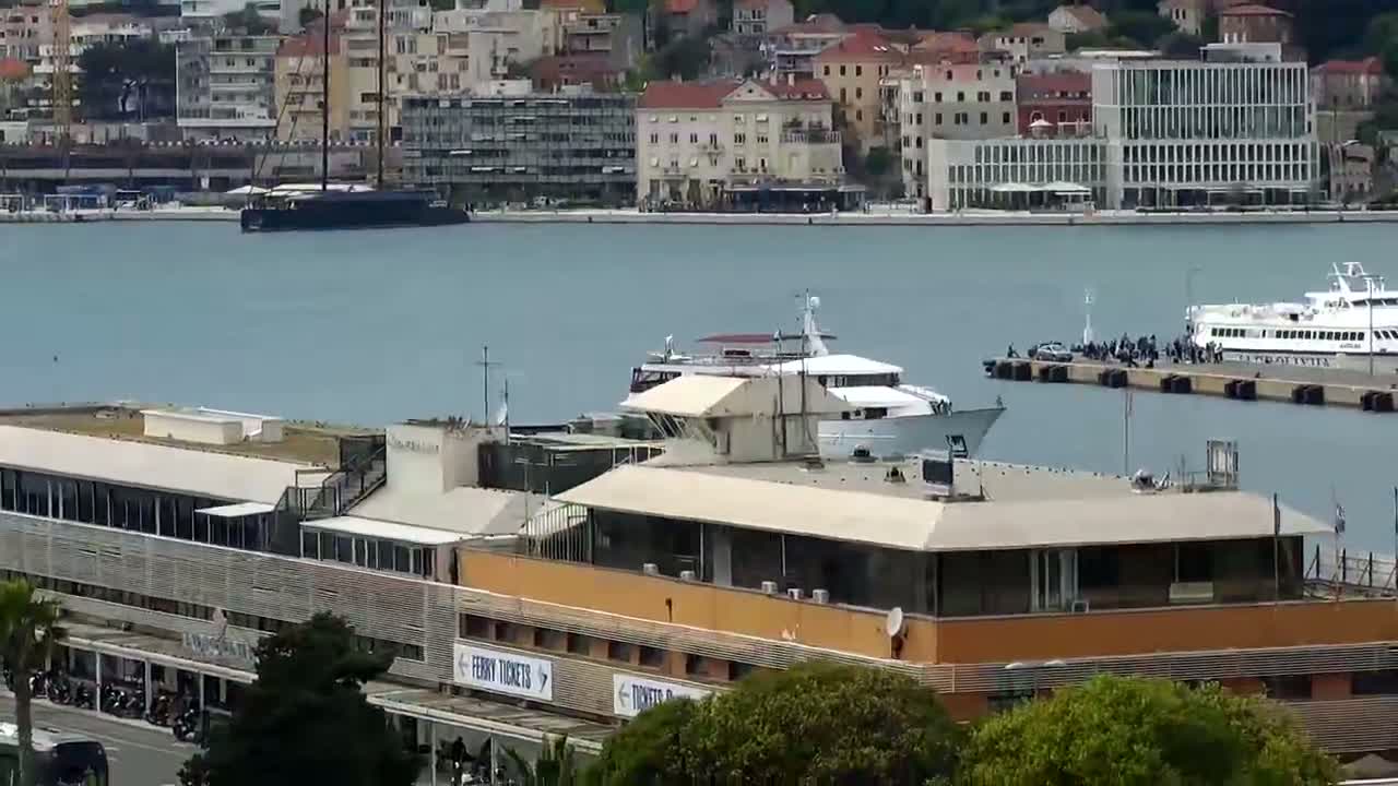 A large building near a calm harbor with several boats, including a yacht and a ferry, in view of a city skyline.