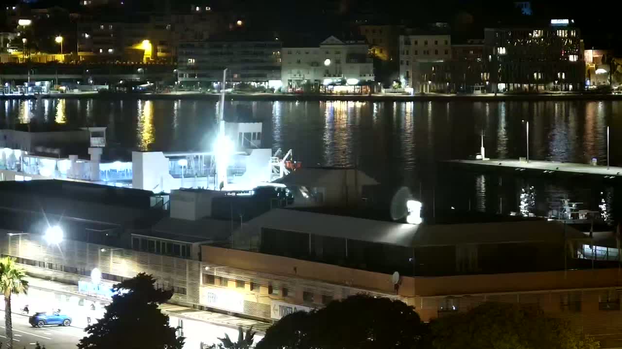 A nighttime view of a harbor with a ferry docked, buildings illuminated along the waterfront, and calm water reflecting city lights.