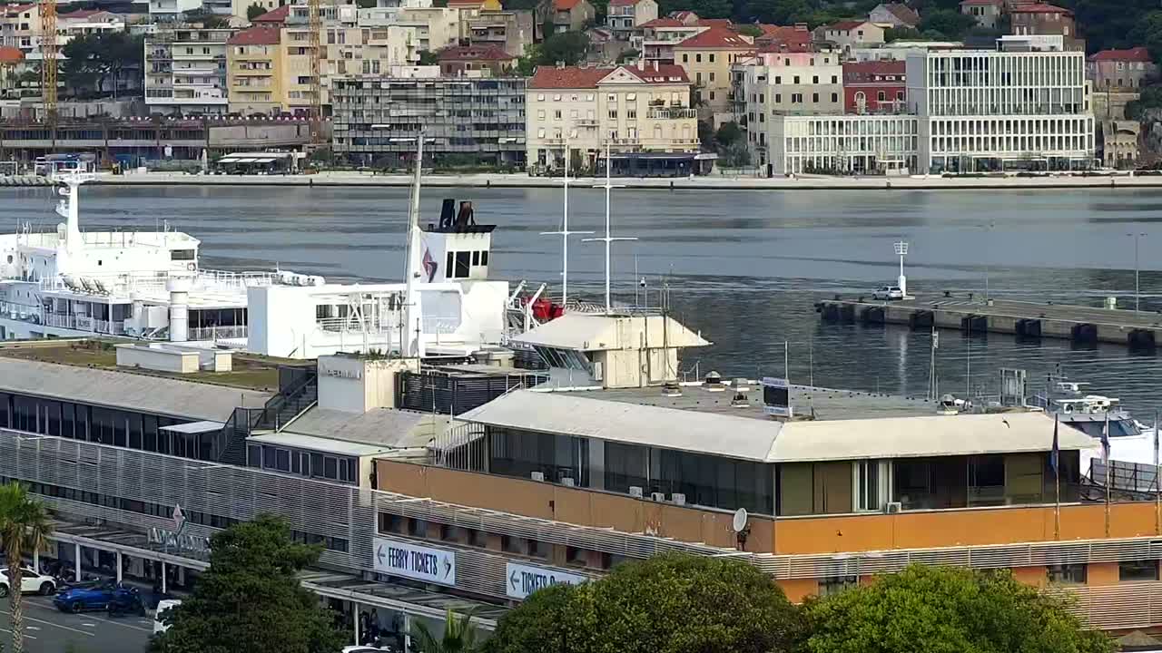 A large white ferry is docked near a multi-story building at a harbor, with calm water and a cityscape in the background on a sunny day.