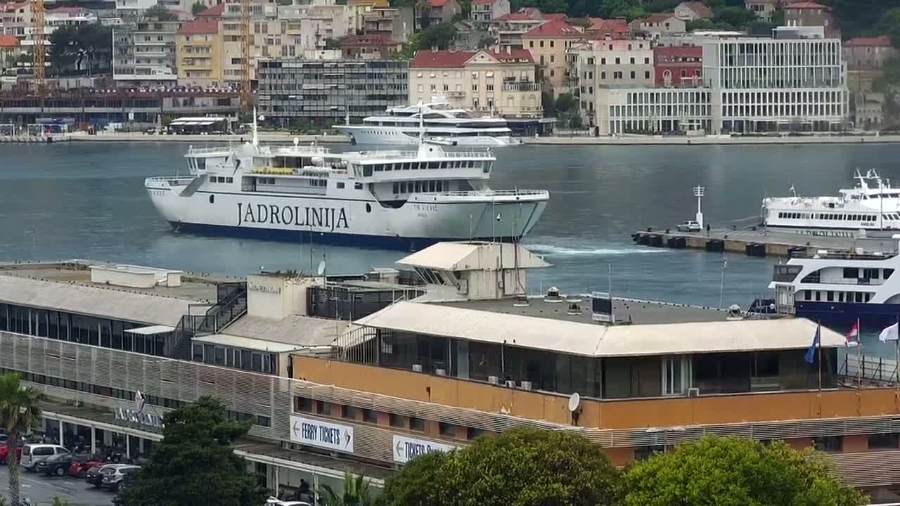 A large ferry departs a harbor in calm water, passing by several smaller vessels and a waterfront building.