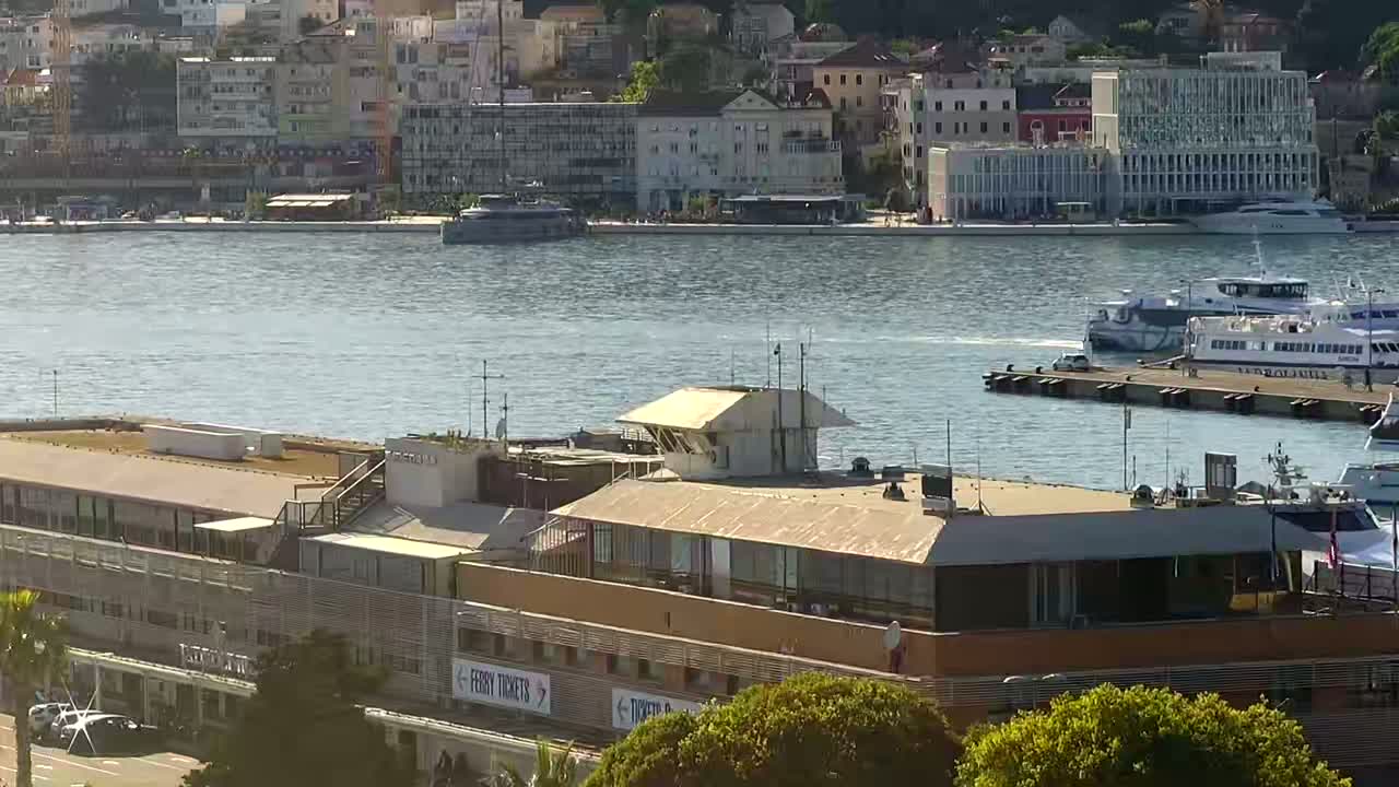 A large ferry terminal building sits on the waterfront, with several ferries docked nearby on a calm, sunny day.