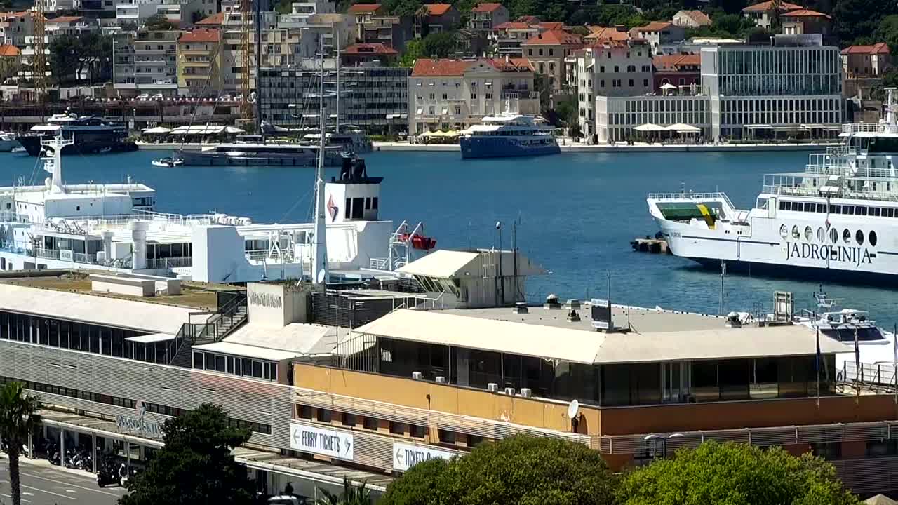 A harbor with several ships docked, including a large white ferry, and a modern building with a flat roof overlooking the water on a sunny day.